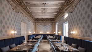A formal dining room with ornate ceiling and wall designs, long tables with chairs, and blue upholstered booths. - Palazzo Castagna (Ghaxaq)