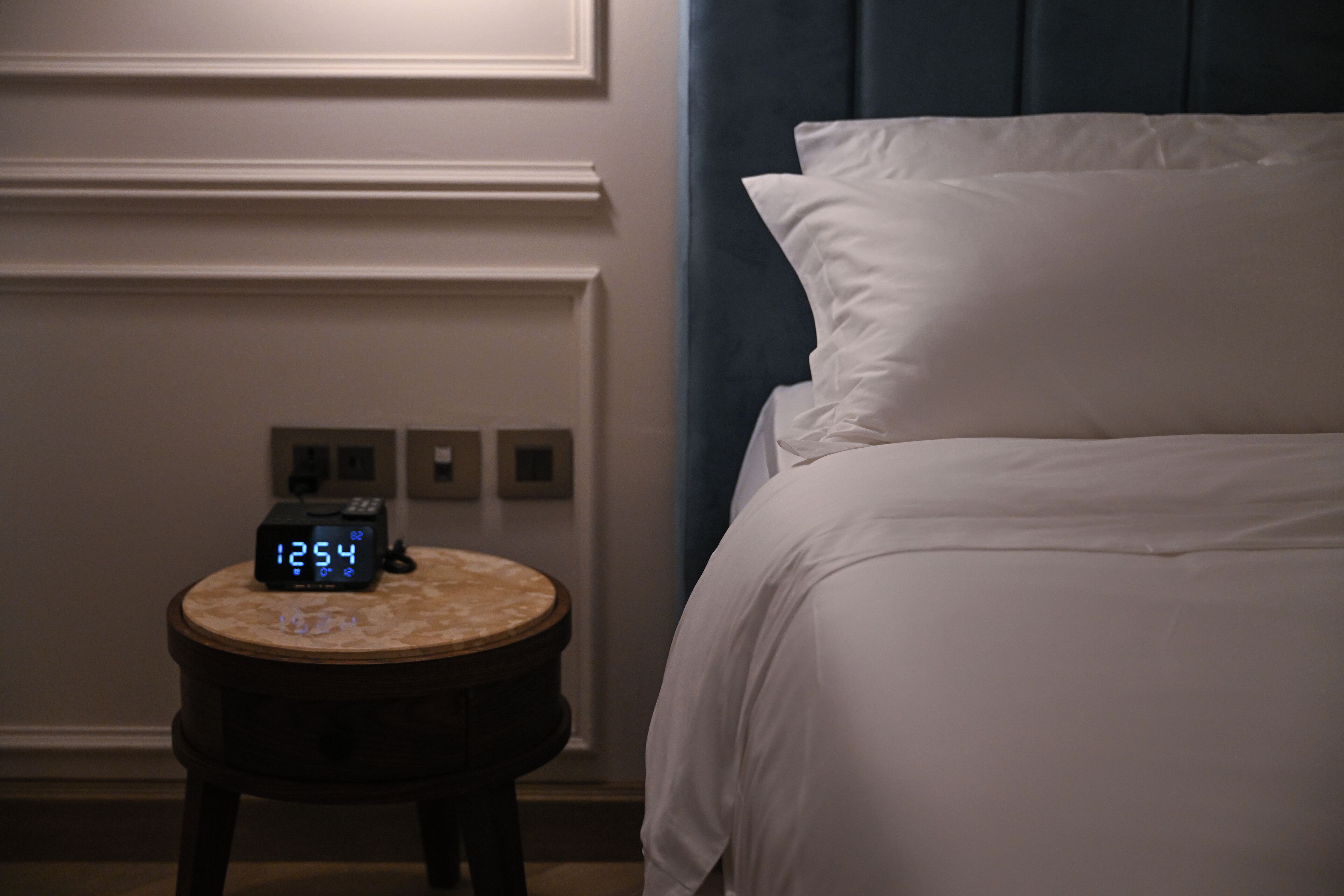 A hotel room with a bed, a round wooden nightstand with a digital clock, and a wall with a light switch and electrical outlets.