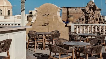 Outdoor seating with wicker chairs and tables, a stone wall with decorative elements, and a view of a building with a dome.