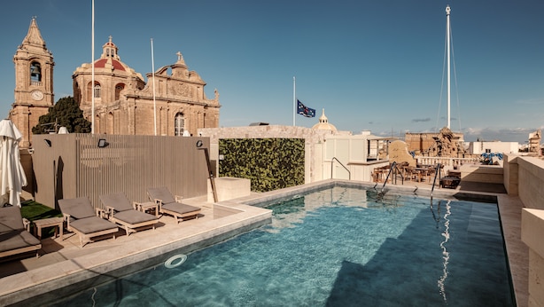 A rooftop pool with lounge chairs, a flag, and a historic building in the background. - Palazzo Castagna (Ghaxaq)