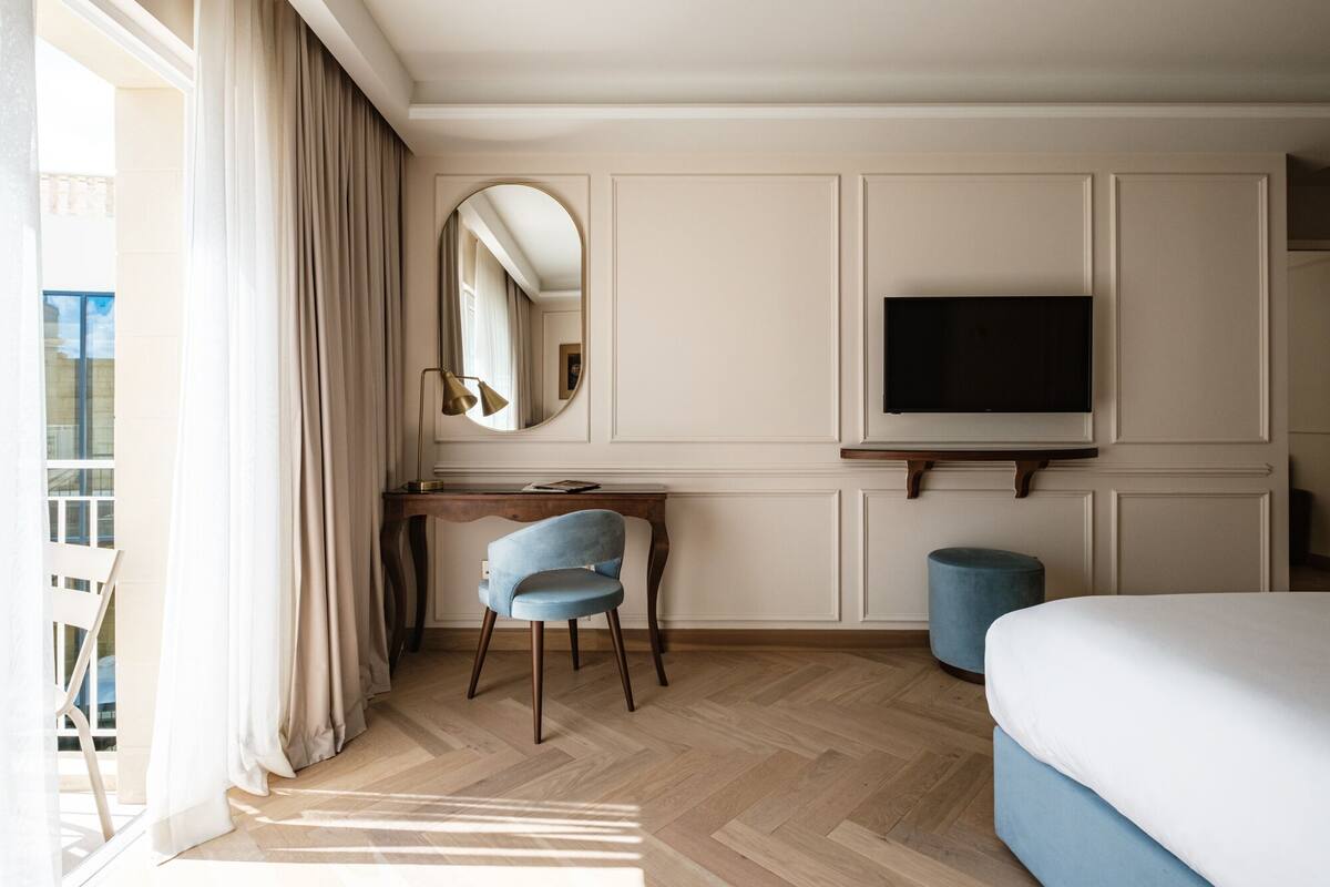 A hotel room with a bed, a desk with a chair, a television, a round mirror, and a window with curtains.