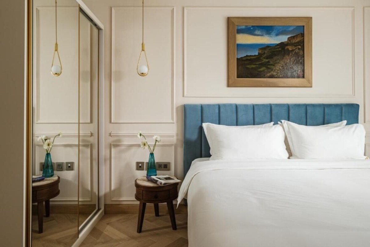 A bedroom with a large bed, a blue upholstered headboard, two bedside tables with vases, and a painting on the wall.