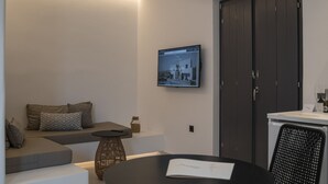 A modern living room with a sofa, a small round table, a flat-screen TV mounted on the wall, and a dark wooden door. - Luxury hospitality, outstanding view (Thira - Santorini)