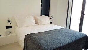 A bedroom with a bed, white pillows, a dark textured blanket, a bedside table with a phone, and a wall-mounted lamp. - Luxury hospitality, outstanding view (Thira - Santorini)