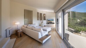 A modern living room with a large sofa, a coffee table, and a view of a mountainous landscape through large windows. - Villa S'Ollastu - Luxury villa with private pool, garden, sea view, near beaches (Chia)