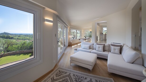 A spacious living room with a large window, a white sofa set, and a patterned rug. - Villa S'Ollastu - Luxury villa with private pool, garden, sea view, near beaches (Chia)