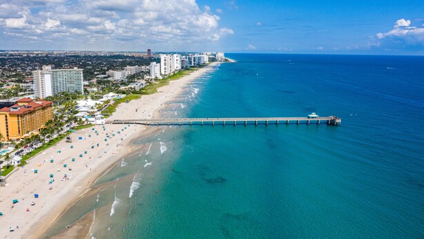 A coastal cityscape with a long pier extending into the sea, a sandy beach lined with umbrellas, and high-rise buildings in the background. - Waterfront Home Steps to Beach (Fort Lauderdale)