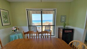 A dining room with a large wooden table, chairs, a small round table with a vase, and a view of the ocean through a large window. - Beachfront - Great Location! (Gulf Shores)