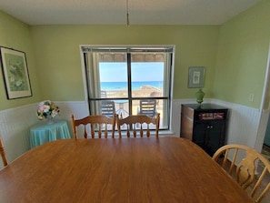 A dining room with a large wooden table, chairs, a small round table with a vase, and a view of the ocean through a large window. - July Dates Discounted!  Beachfront (Gulf Shores)