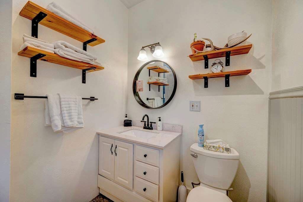 Une salle de bain avec des étagères en bois où sont rangées les serviettes, un miroir rond, un lavabo avec un robinet et des toilettes.