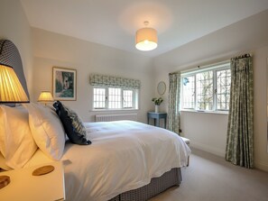 Quarto com cama, luminária de cabeceira, janela com cortinas, radiador e uma pequena mesa com um vaso.