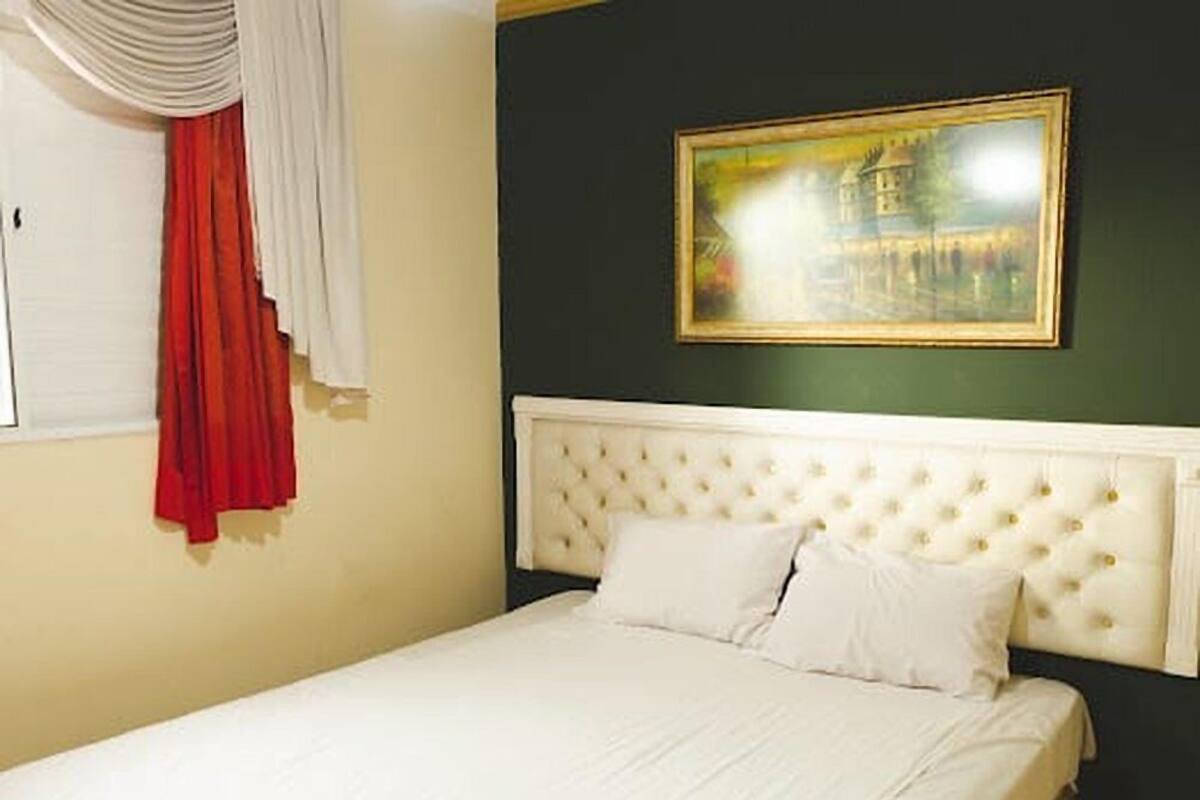 A bedroom with a white bed, a tufted headboard, a painting on the wall, and a red curtain.
