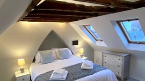 Un dormitorio con techo inclinado, una cama con ropa blanca, dos mesitas de noche con lámparas, un aparador y un tragaluz.