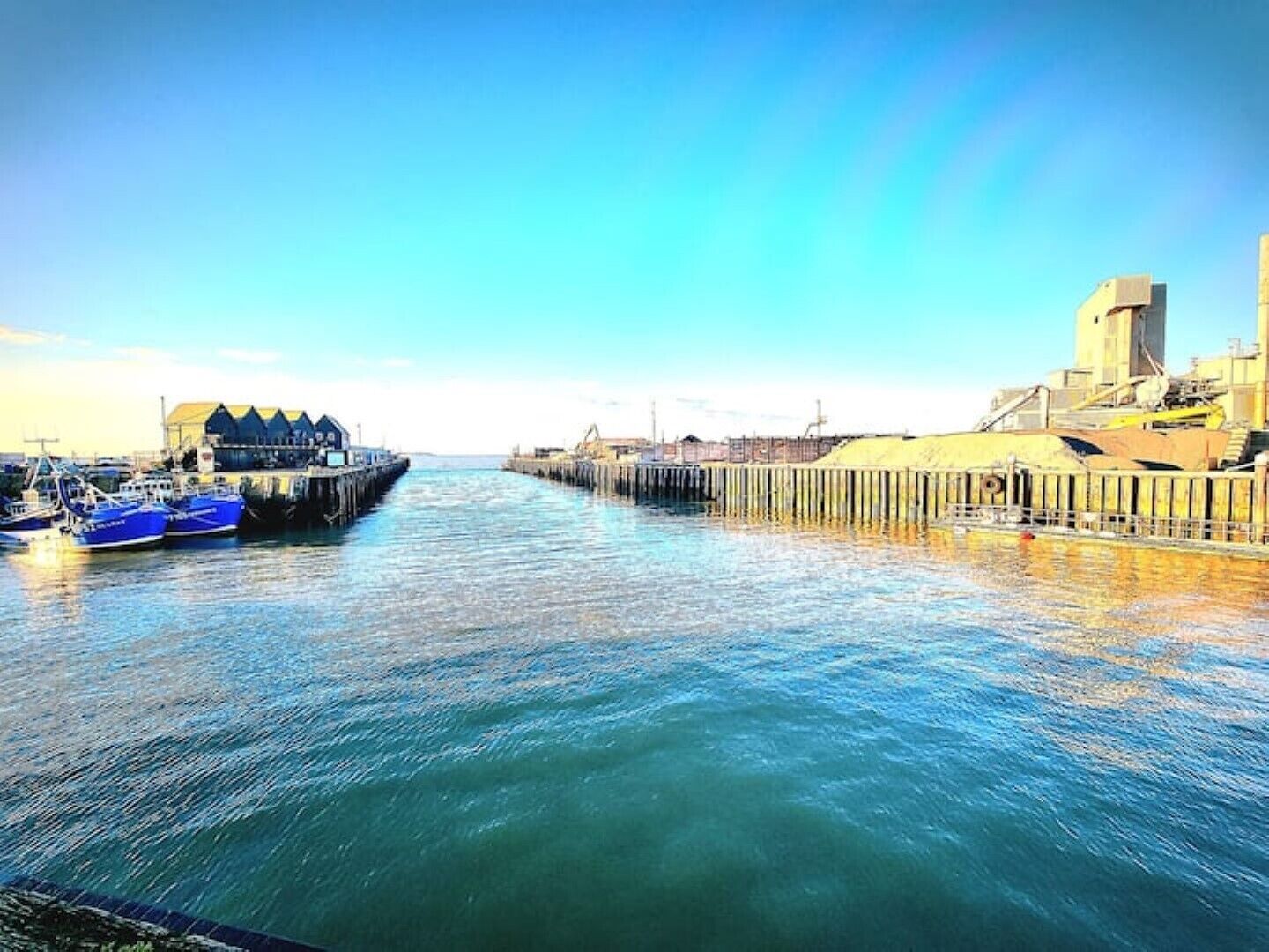 Una escena frente al agua con barcos amarrados en un muelle, un edificio con un techo distintivo y una gran estructura industrial a la derecha.