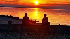 Dos siluetas de personas sentadas en un banco mirando al atardecer sobre un cuerpo de agua.