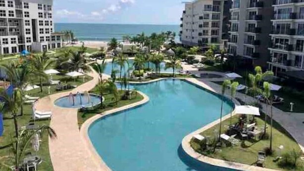 Resort com uma piscina grande em curva, palmeiras e vista para a praia.