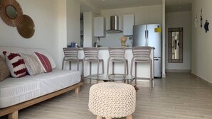 Sala moderna com sofá, puf e poltronas, cozinha com geladeira e área de jantar.