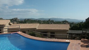 Una piscina con plataforma de baldosas, un edificio con techo de tejas y un paisaje montañoso al fondo.