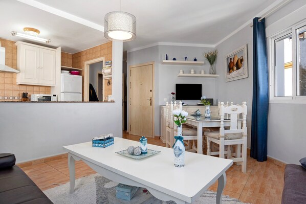 A modern kitchen with white cabinets, a microwave, and a refrigerator. There is a dining area with a white table, chairs, and a vase with flowers. A television is mounted on the wall above the dining table.