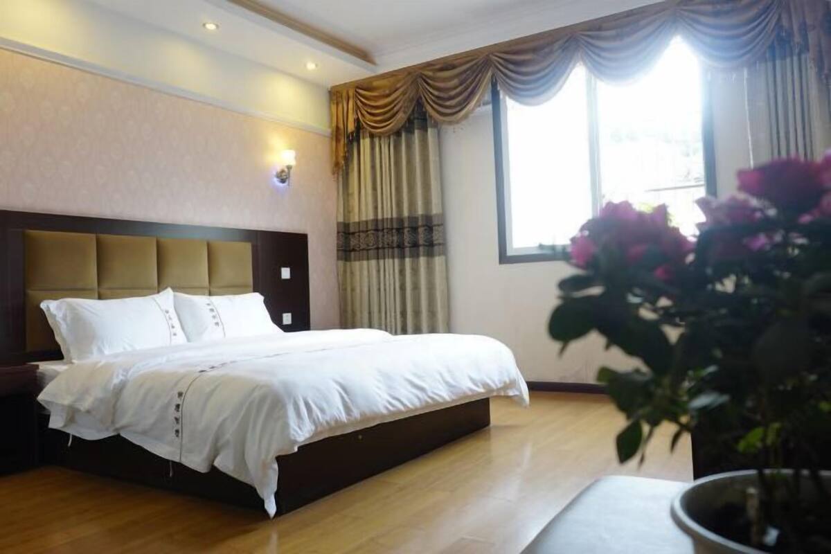 A hotel room with a large bed, a headboard, a potted plant, and a window with curtains.