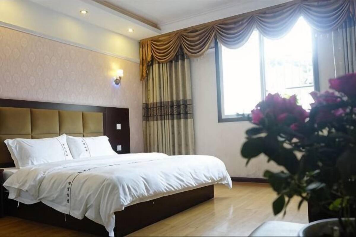 A hotel room with a large bed, a headboard, a window with curtains, and a vase with flowers.