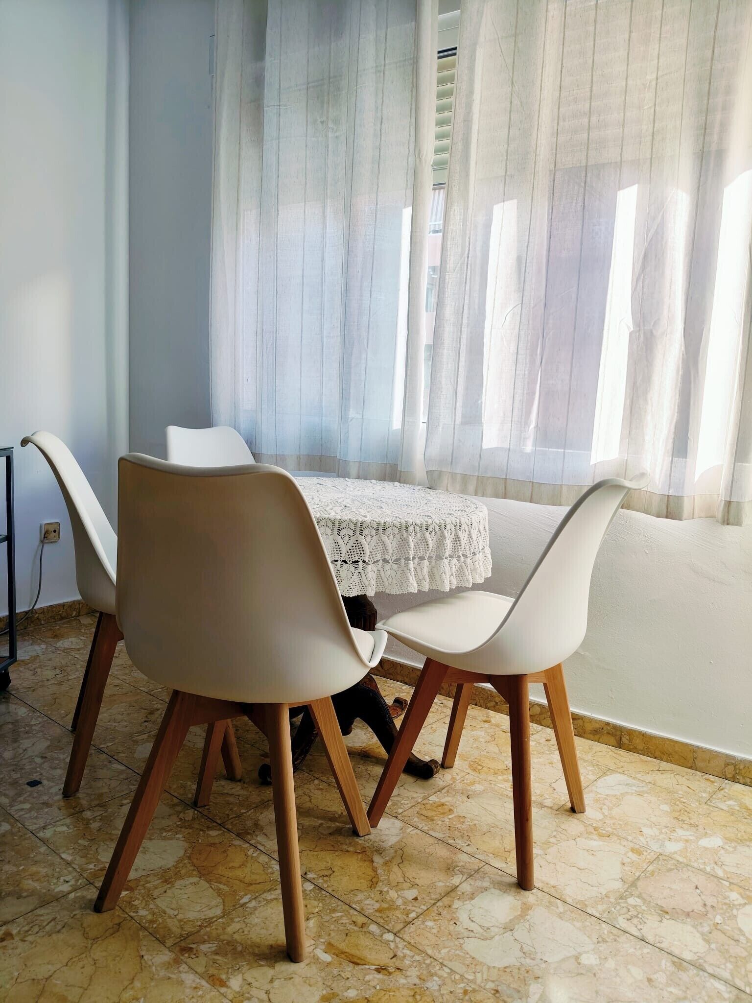 Un coin repas avec une table et des chaises, une fenêtre ornée de rideaux légers et un sol carrelé.