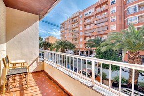 A balcony with a chair, a railing, and a view of a multi-story building with balconies and palm trees.