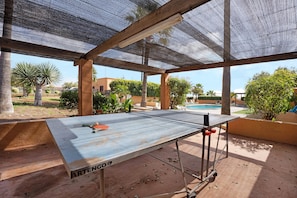 Une table de ping-pong extérieure, avec un filet, située dans un espace ombragé d’où l’on peut admirer une piscine et des palmiers.