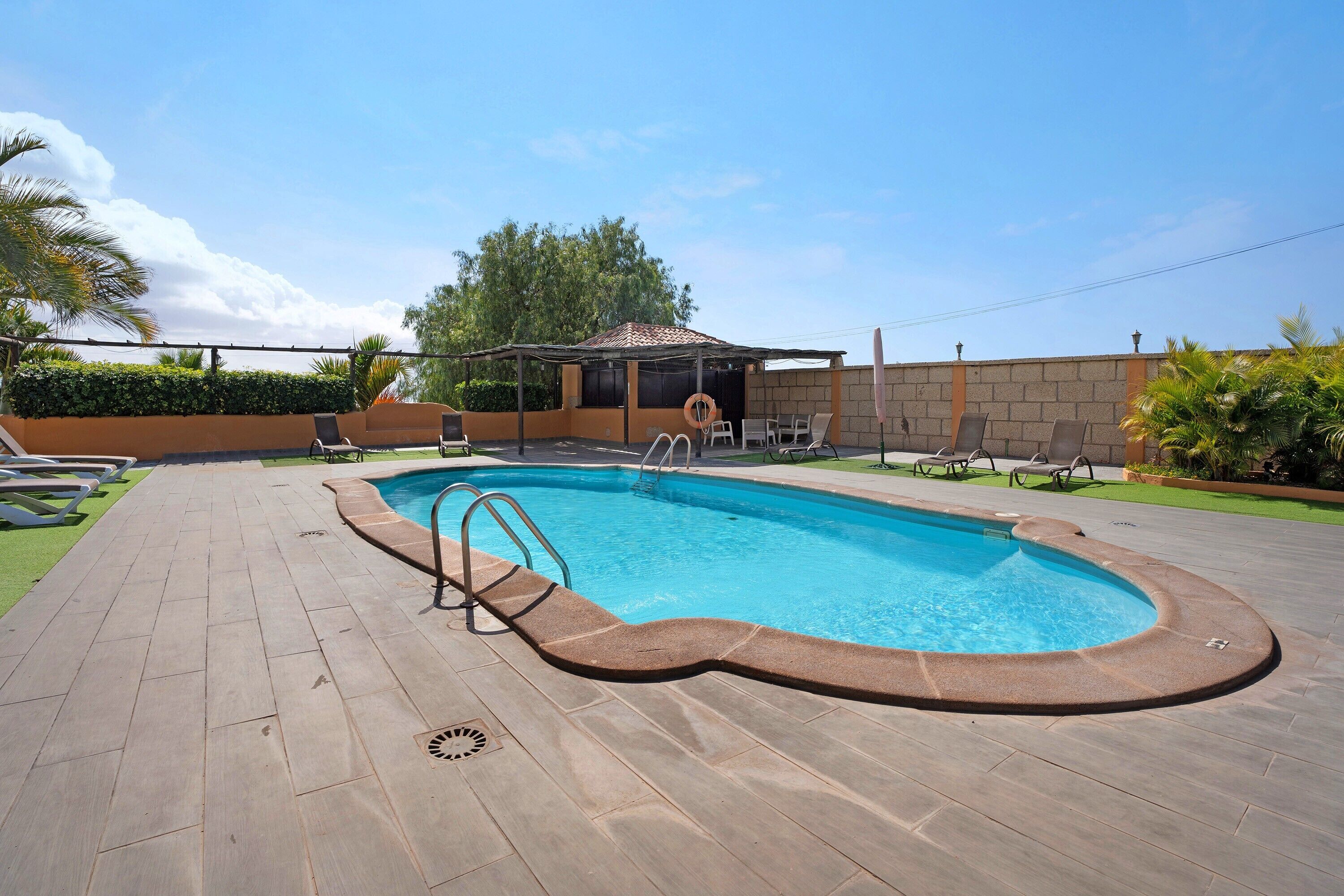 A swimming pool with a wooden deck, lounge chairs, and a covered seating area.