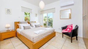 A bedroom with a large bed, a nightstand, a chair, a window with a view of palm trees, and a ceiling fan. - Villa Sophia - 3 bed villa with private pool, 2 min walk to beach and internet (Paralimni)