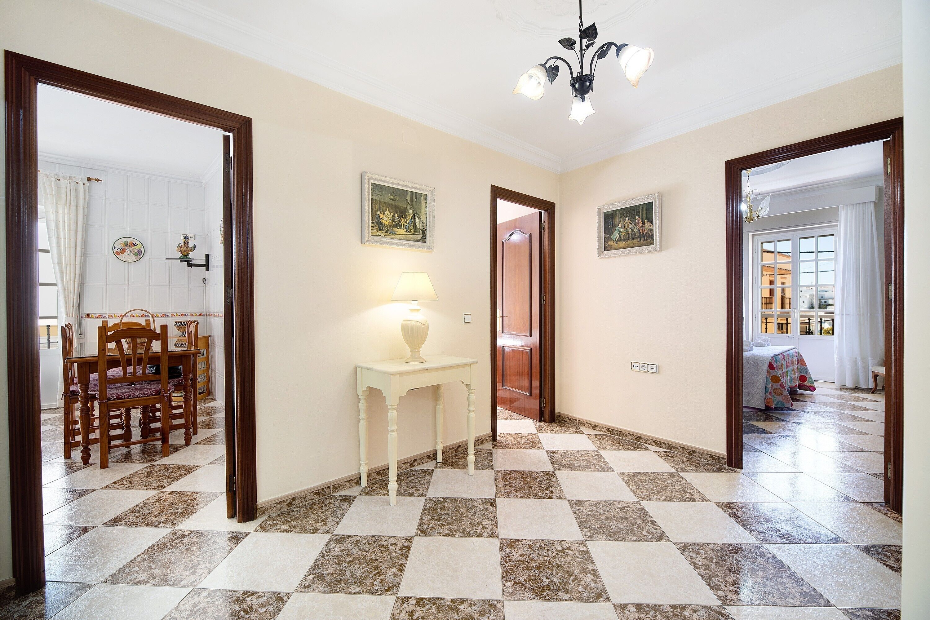A hallway with a checkered floor, a dining area with chairs and a table, and a bed with a colorful bedspread in the background.