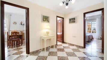 A hallway with a checkered floor, a dining area with chairs and a table, and a bed with a colorful bedspread in the background.
