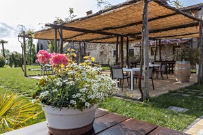 Un jardín con una mesa de madera, una planta en maceta con flores y una zona de estar al aire libre cubierta con mesas y sillas.