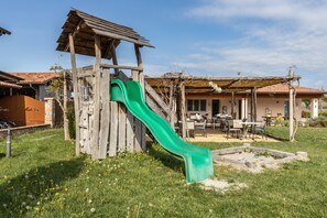 Una estructura de juego de madera con un tobogán verde, un patio con mesas y sillas, y un edificio con techo de tejas al fondo.