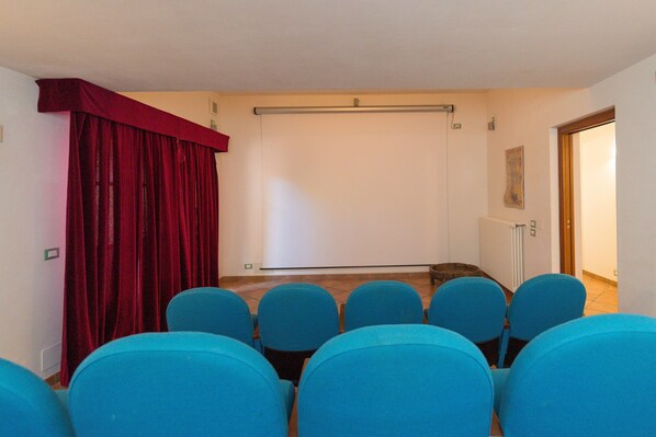 Un salón de conferencias con una pantalla de proyección blanca, cortinas rojas y sillas azules.