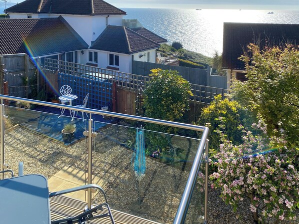 A balcony with a view of the sea, a garden with potted plants, and a white house with a tiled roof.