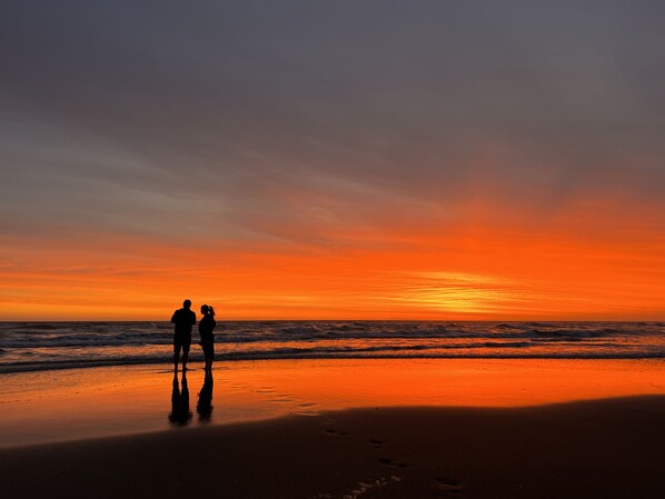 Eine Strandansicht, zwei Personen sind vor einem Sonnenuntergang im Schatten zu sehen.