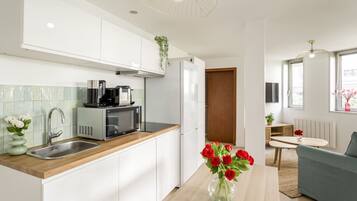 Cocina moderna con armarios blancos, un lavamanos de acero inoxidable y un microondas. Hay un comedor con una mesa y un sofá azul.