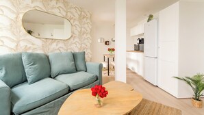 Un salon moderne avec un canapé, une table basse ornée d’un vase de fleurs et un coin cuisine doté de meubles blancs et d’appareils électroménagers.