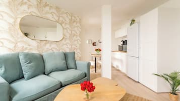 Una sala moderna con un sofá, una mesa de centro con un florero, y un área de cocina con armarios y electrodomésticos blancos.