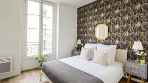 A bedroom with a large bed, a window with a view, a patterned wallpaper, and a bedside table with a lamp. - Family Suite - Opera - Caumartin II (Paris)