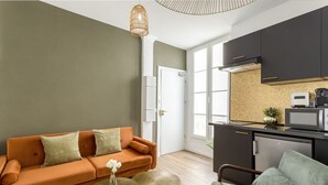 A modern living room with a sofa, coffee table, and chairs, a kitchen with dark cabinets and a microwave, and a large window providing natural light. - Family Suite - Opera - Caumartin II (Paris)