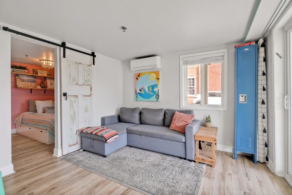 A modern living room with a grey sectional sofa, a small wooden side table, and a grey patterned rug. There is a blue locker against the wall and a large window with a view of the outside.