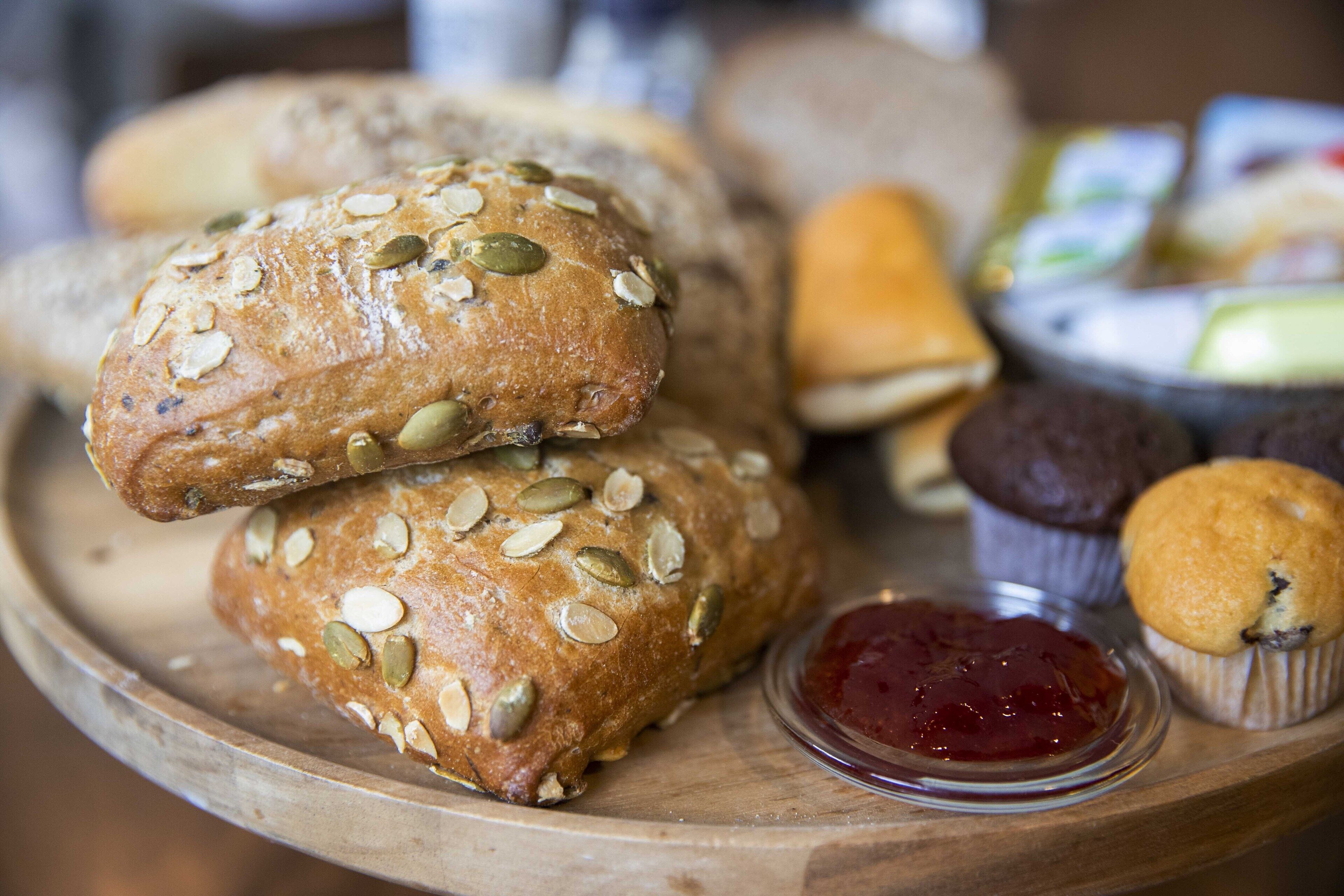 A wooden board with various breads and muffins, including pumpkin seed and oat breads, alongside a small container of jam.