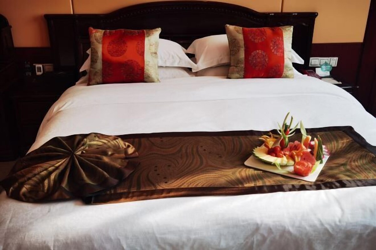 A neatly made bed with white linens, two decorative pillows, a wooden tray with a fruit salad, and a brown decorative pillow.