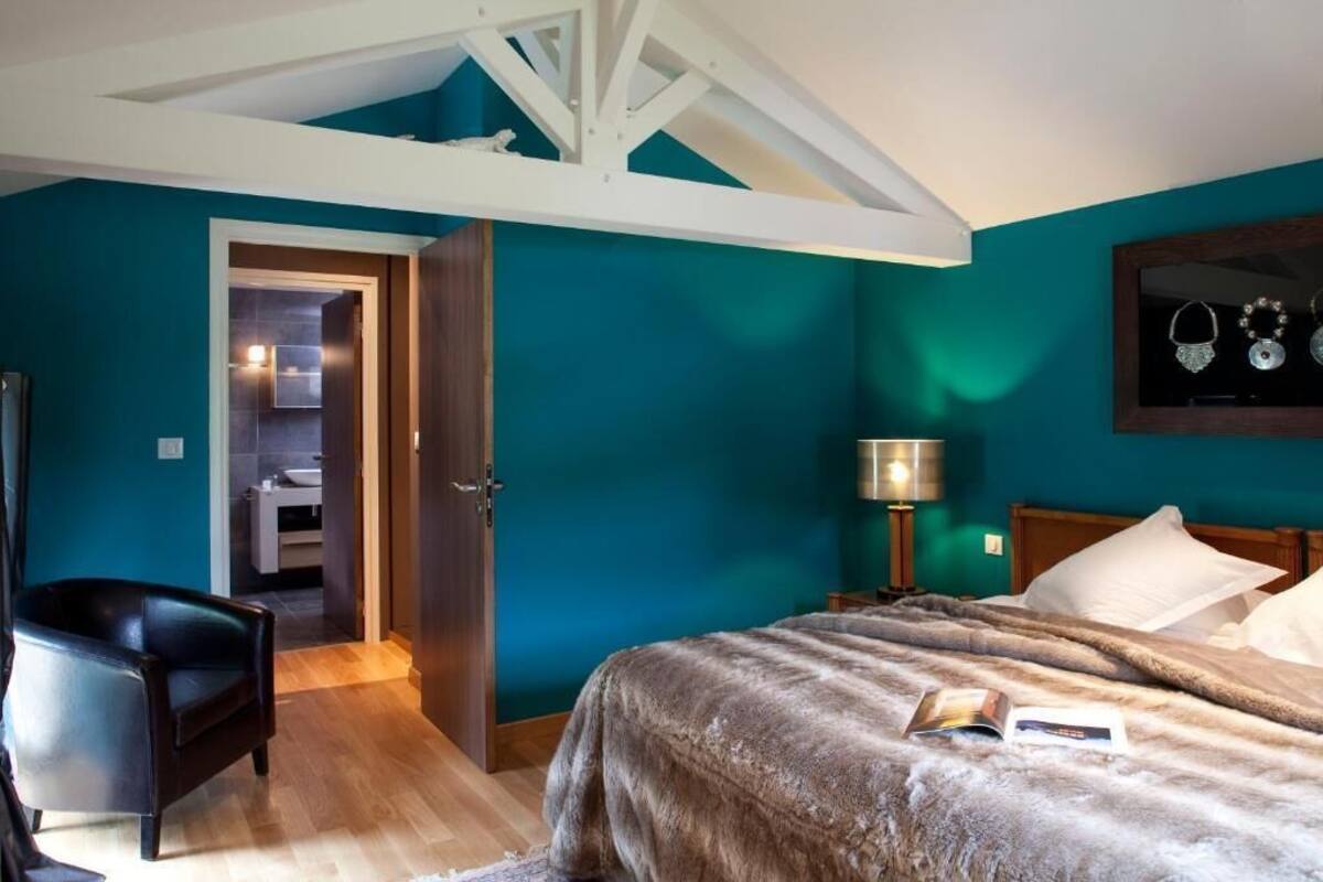 A bedroom with teal walls, a wooden ceiling, a bed with a fluffy blanket, a black chair, a bedside table with a lamp, and a framed jewelry display.