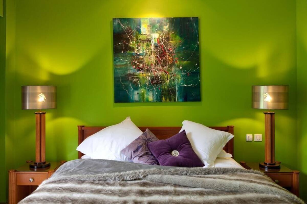 A hotel room with a green wall, a bed with a grey and white striped blanket, two bedside lamps, and a colorful abstract painting above the bed.