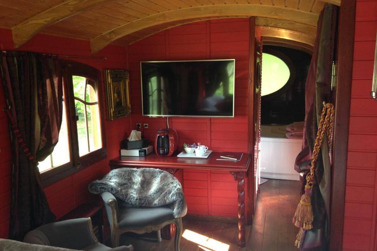 A cozy room with a red wall, a flat-screen TV mounted above a wooden desk, a chair, and a window with curtains.