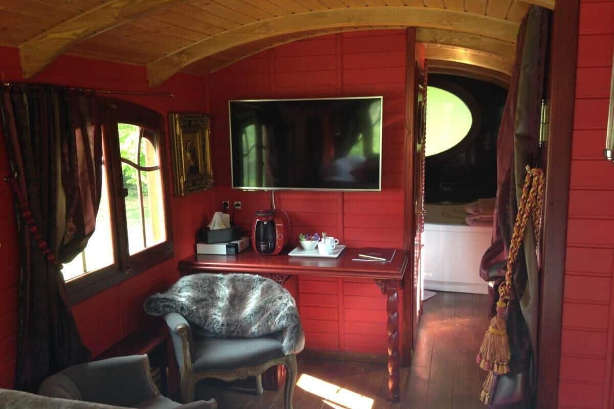 Una habitación acogedora con una pared roja, un televisor de pantalla plana sobre un escritorio de madera, una silla y una ventana con cortinas.
