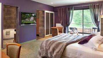 A hotel room with a large bed, a TV mounted on the wall, a seating area with a chair and table, and a view of the outdoors through the window.